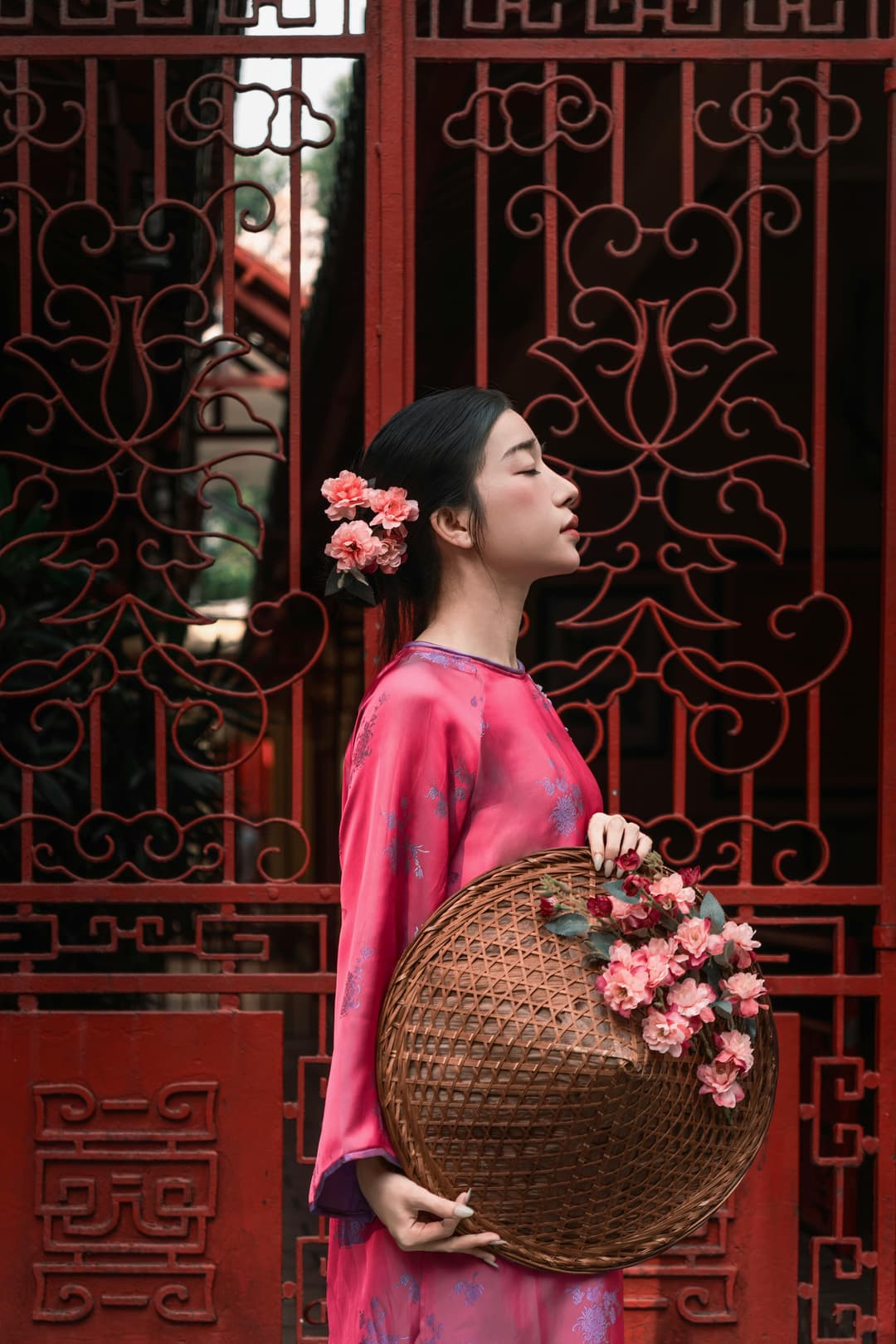A woman in a pink traditional dress stands in profile before an ornate red gate, holding a woven basket decorated with pink flowers, with matching flowers in her hair.